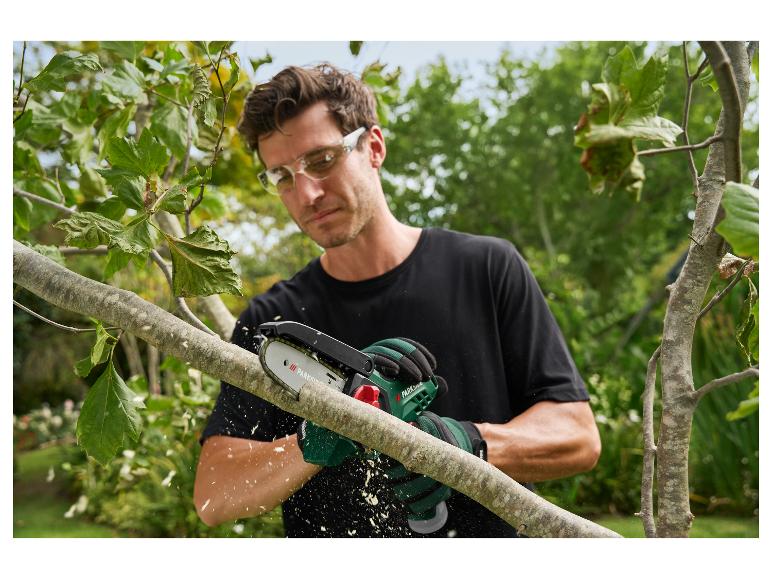 Mann schneidet einen Ast mit einer Kettensäge, trägt Schutzbrille und Handschuhe.
