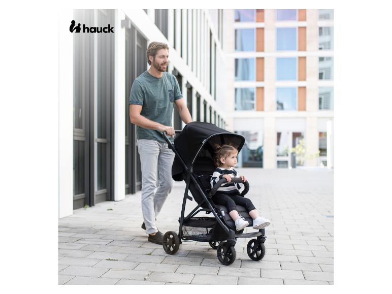 Mann schiebt einen schwarzen Hauck Kinderwagen mit einem Kind darin, draußen.
