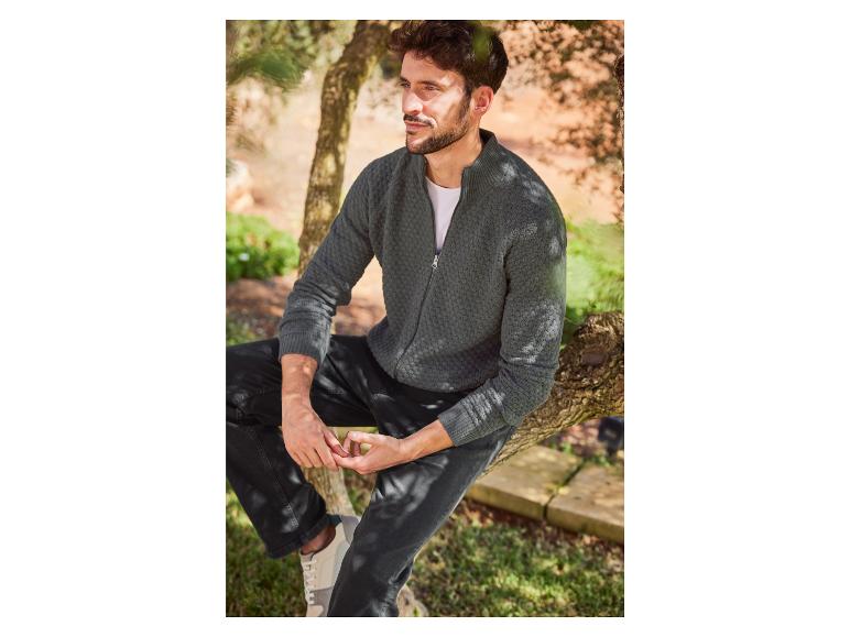 Mann in einem grauen Reißverschluss-Pullover, schwarzen Hosen und weißen Sneakern.