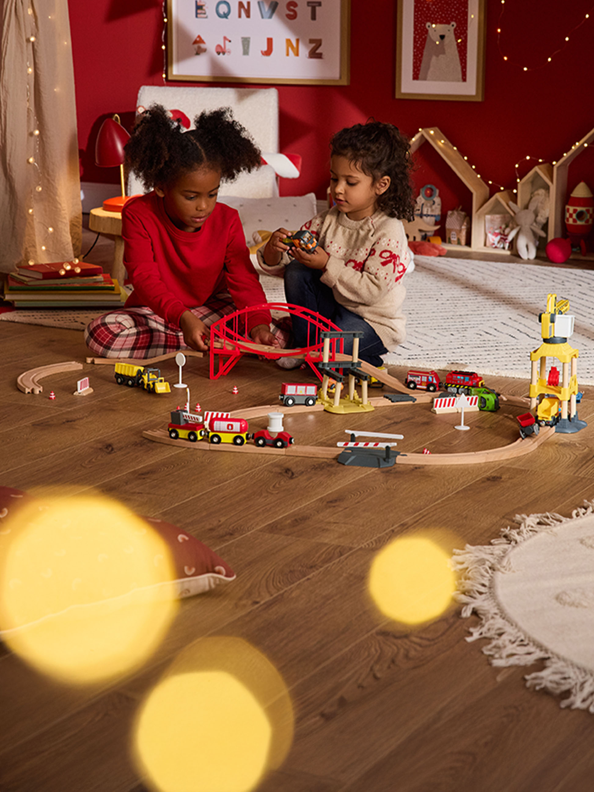 Zwei Kinder spielen mit einer Holzeisenbahn und Fahrzeugen auf einem Holzboden.