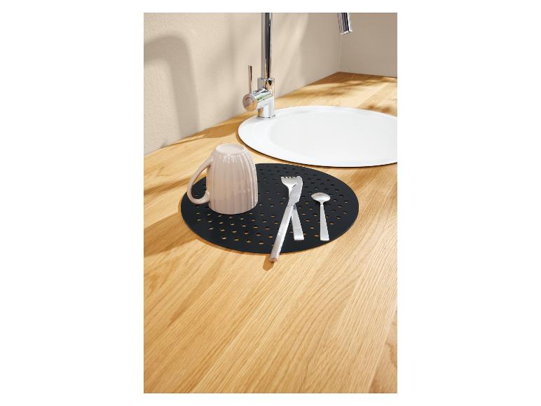 Eine schwarze, gelochte Trocknungsmatte liegt auf einer hellen Holzarbeitsplatte und hält eine Tasse und Besteck in der Nähe eines weißen Spülbeckens.