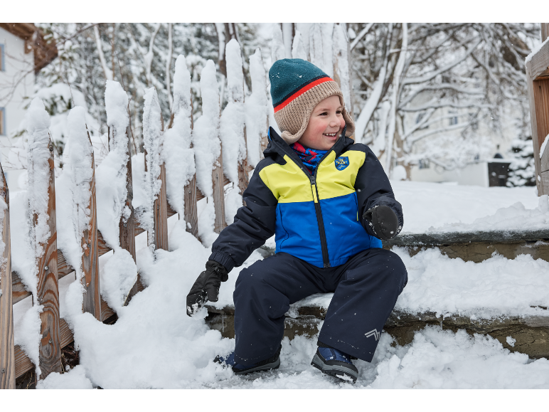 Lächelndes Kind in Skijacke und Schneehose sitzt auf verschneiten Stufen.