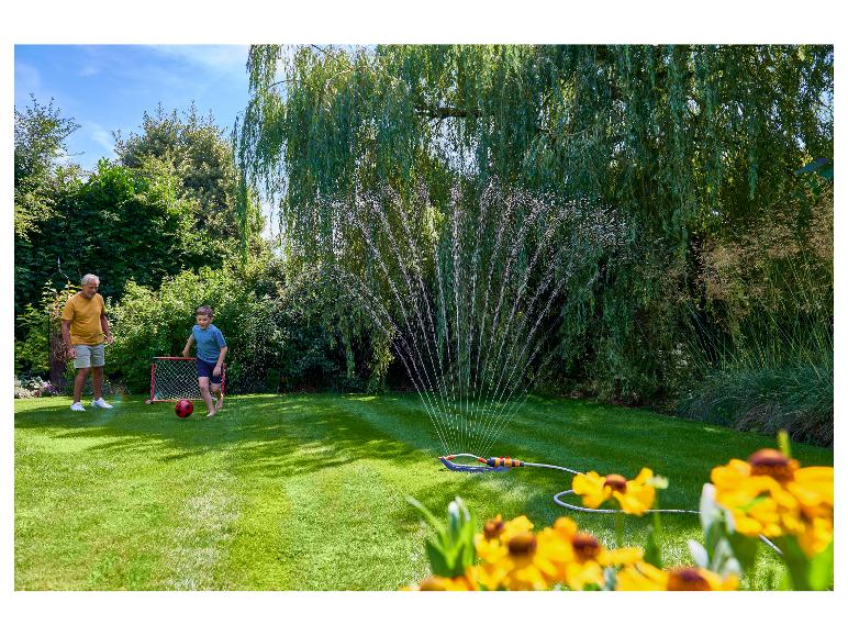 Großvater und Kind spielen Fußball im Garten, mit einem Rasensprenger im Hintergrund.