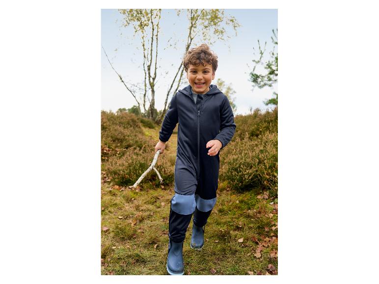 Lächelnder Junge in einem dunkelblauen wasserdichten Overall und Stiefeln, der einen Stock im Wald hält.