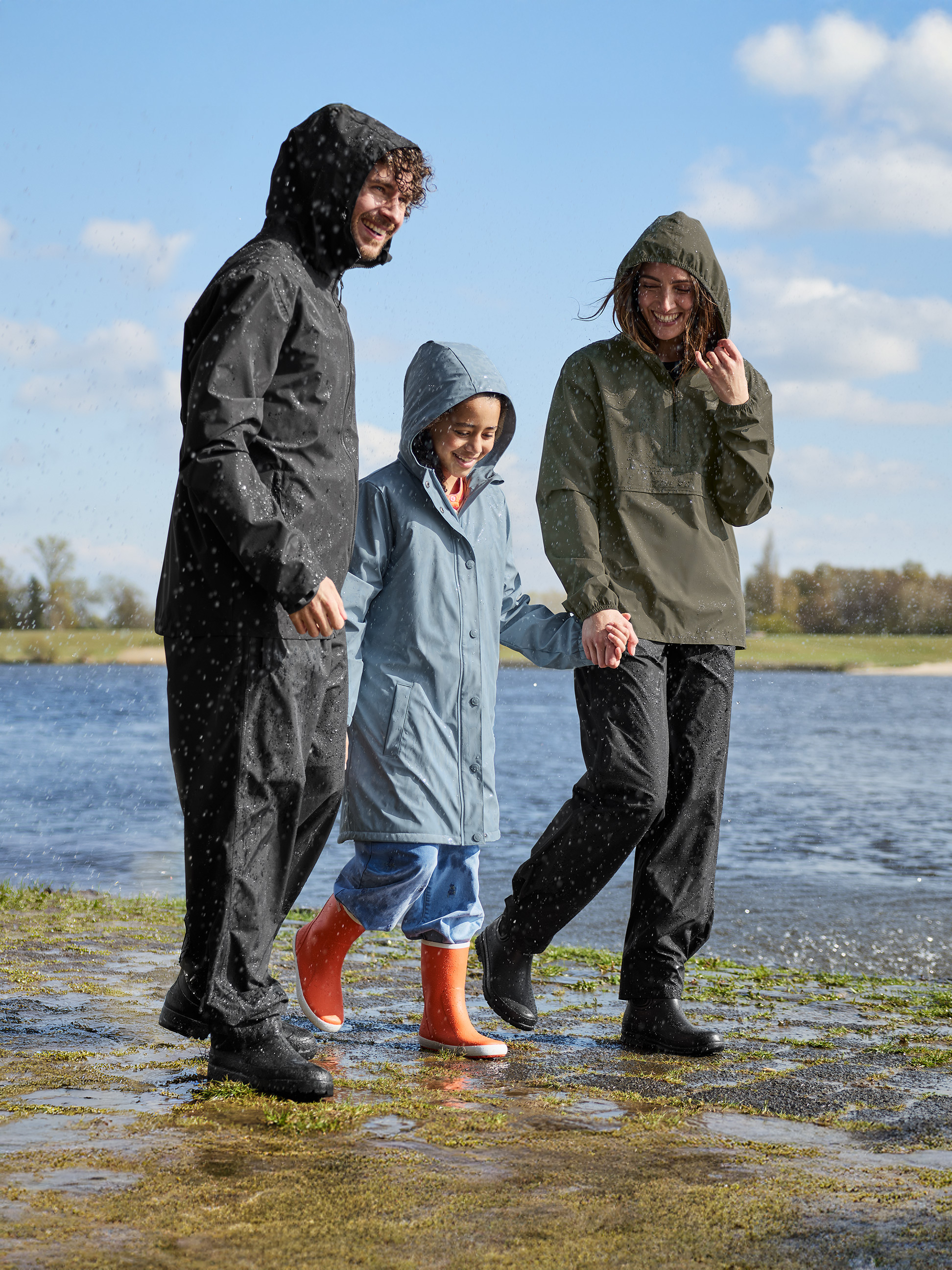 Lächelnde Familie in Regenmänteln und Stiefeln, die am Wasser spaziert.