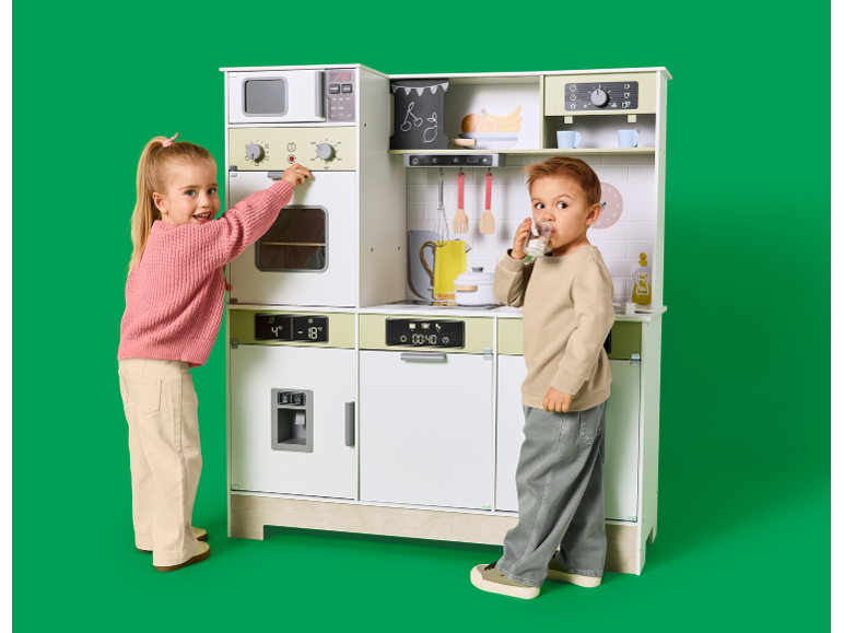 Zwei Kinder spielen mit einer großen Kinderküche aus Holz vor grünem Hintergrund.