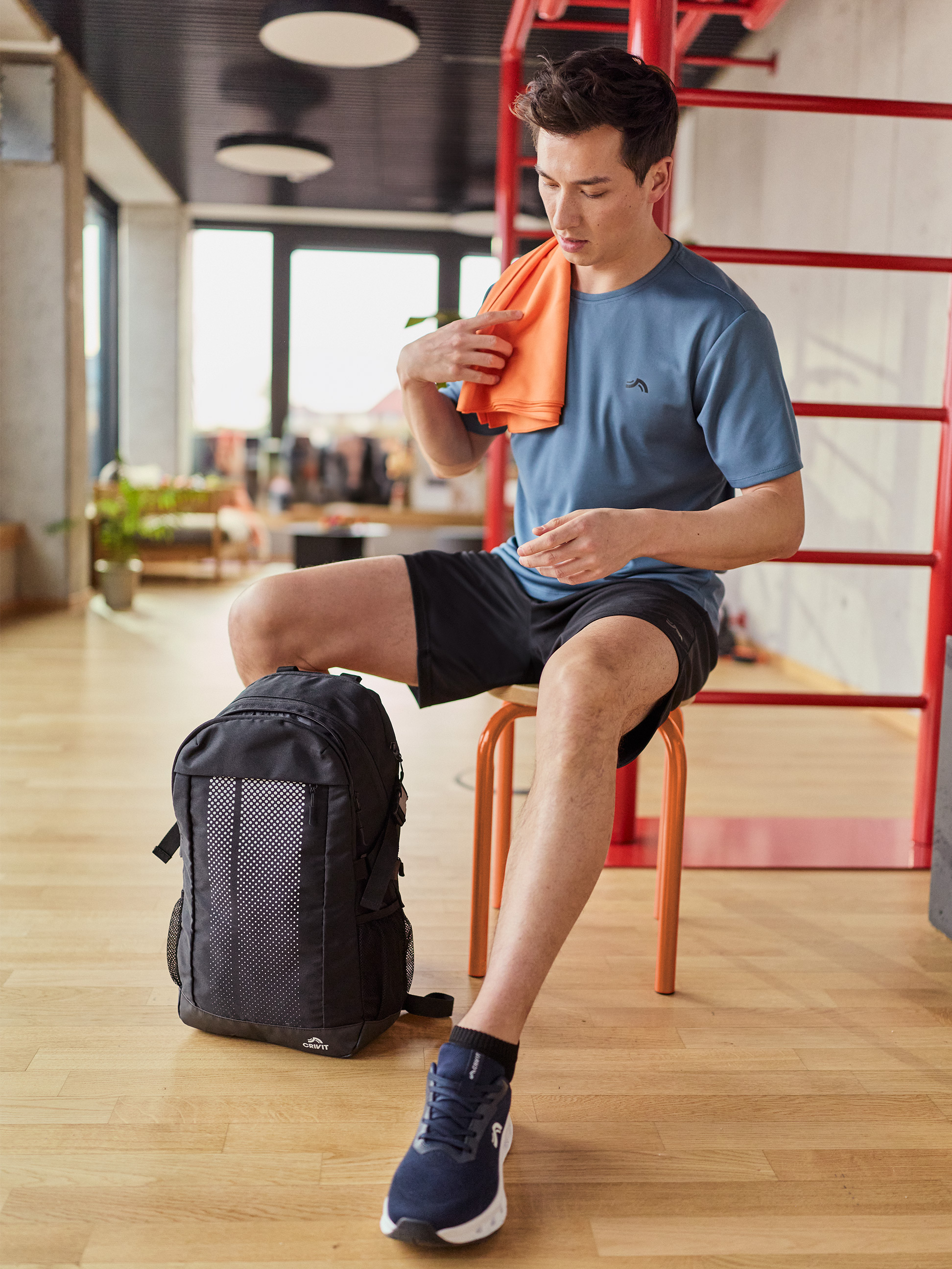 Mann in Sport-T-Shirt und Shorts, trocknet sich mit orangefarbenem Handtuch, mit Rucksack daneben.