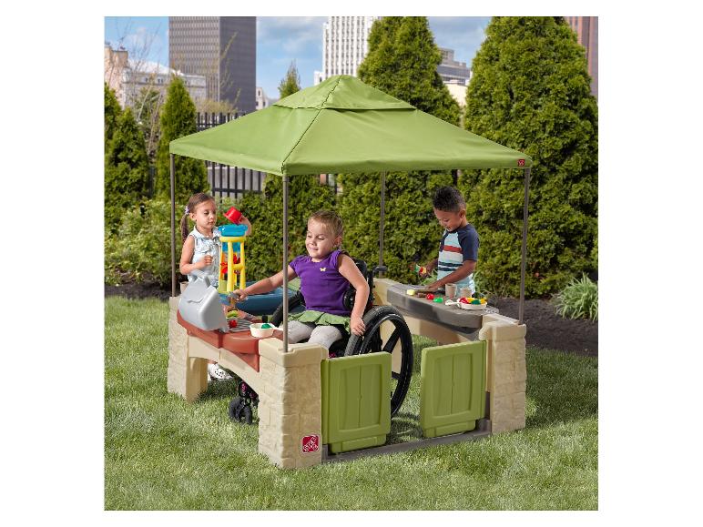 Kinder spielen mit einer Outdoor-Spielküche mit Überdachung, darunter ein Mädchen im Rollstuhl.