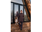 Frau in brauner Jacke und Hose mit Hund vor einem Holzhaus.