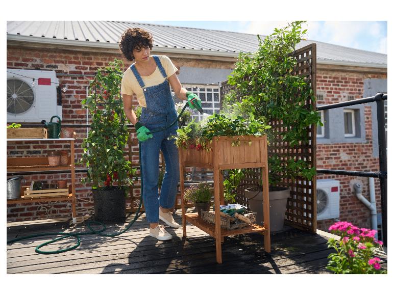 Eine Person gießt Erdbeerpflanzen in einem erhöhten Holzbeet auf einer Dachterrasse.