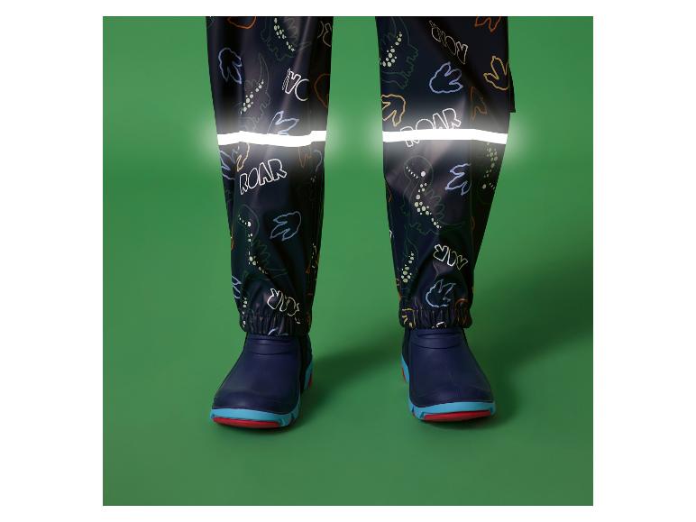 Kinder-Regenhose mit Dinosaurier-Print und reflektierenden Streifen, kombiniert mit blauen Stiefeln.