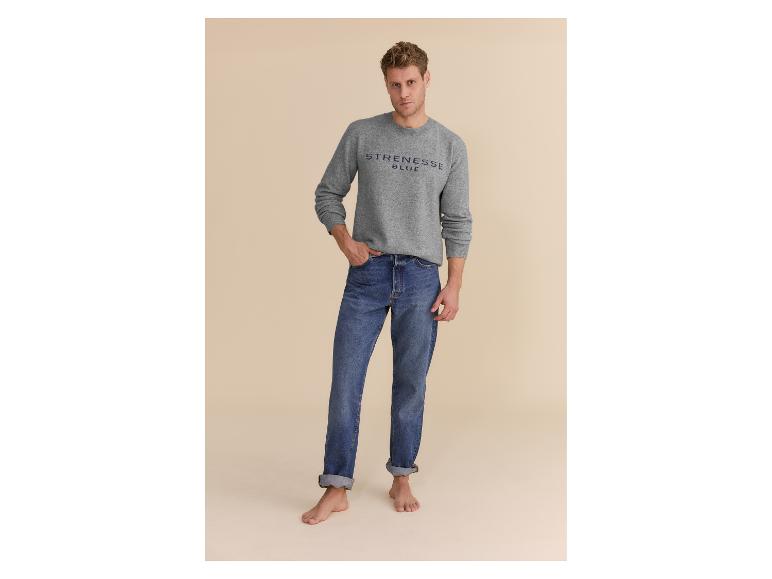 Mann in grauem Pullover mit Logo und blauen Jeans mit umgekrempelten Hosenbeinen