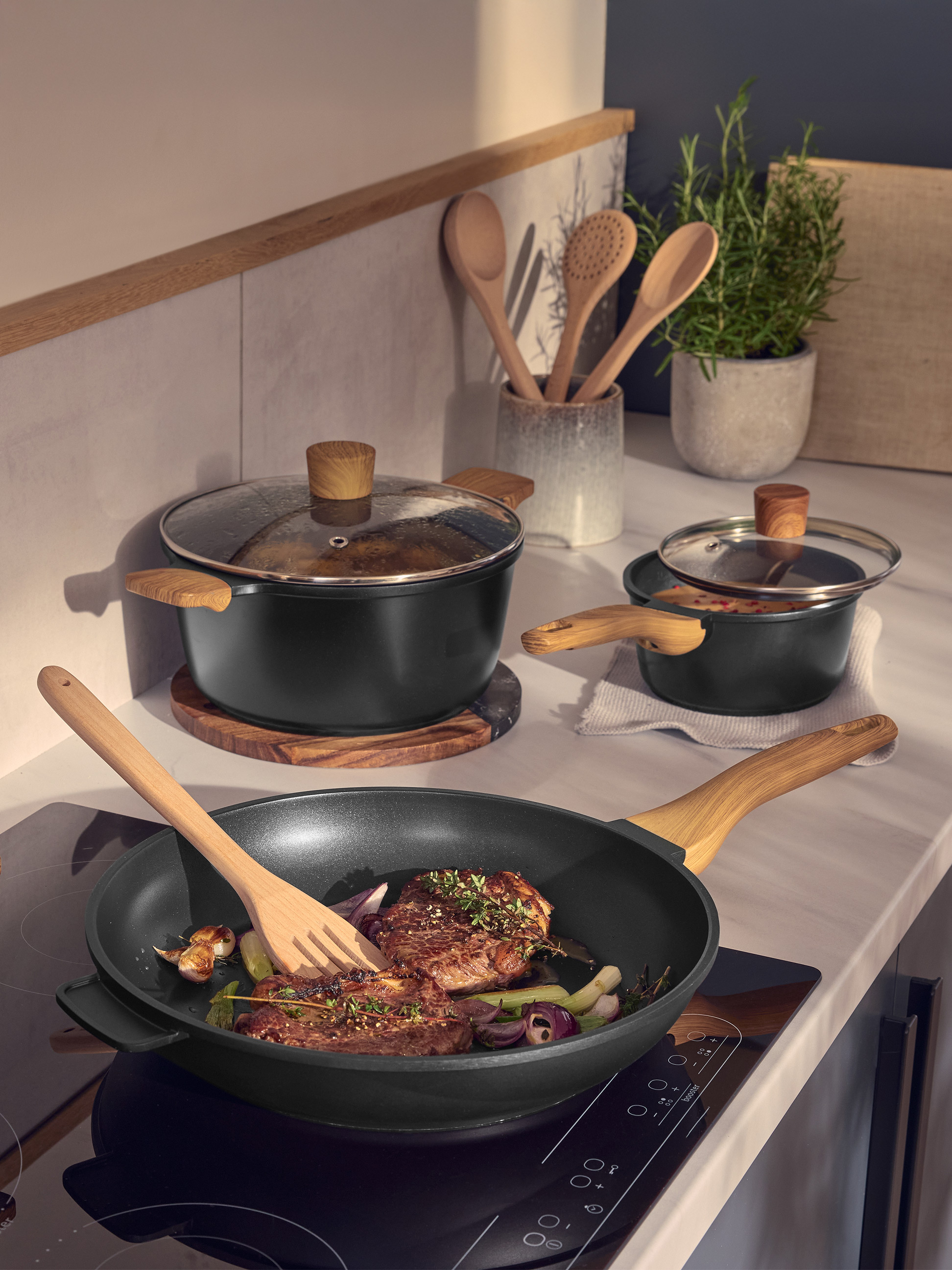 Schwarze Pfannen und Töpfe mit Holzgriffen, die Fleisch und Gemüse in einer modernen Küche zubereiten.