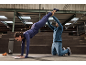 Frau in blauem Sportoutfit macht einen Handstand auf einer Matte mit Hilfe eines Trainers.