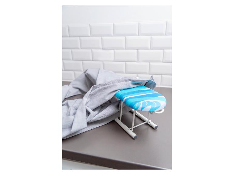 Ein Mini-Bügelbrett mit einer blau-weiß gestreiften Abdeckung steht auf einer grauen Arbeitsplatte, über die ein graues Hemd gelegt ist.