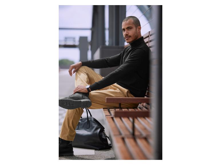 Ein Mann sitzt auf einer Bank mit einem schwarzen Pullover, beige Hosen und schwarzen Stiefeln.