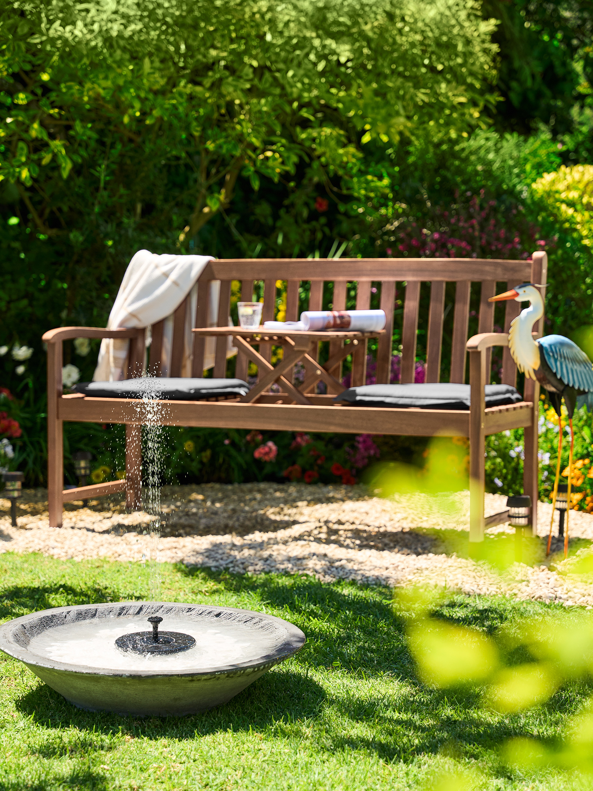 Holzgartenbank mit kleinem Tisch, Kissen und einem dekorativen Reiher in einem sonnigen Garten.