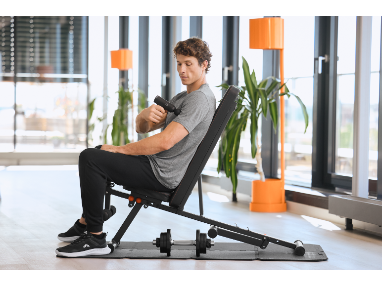 Mann benutzt eine Massagepistole auf einer Hantelbank in einem modernen Fitnessstudio.