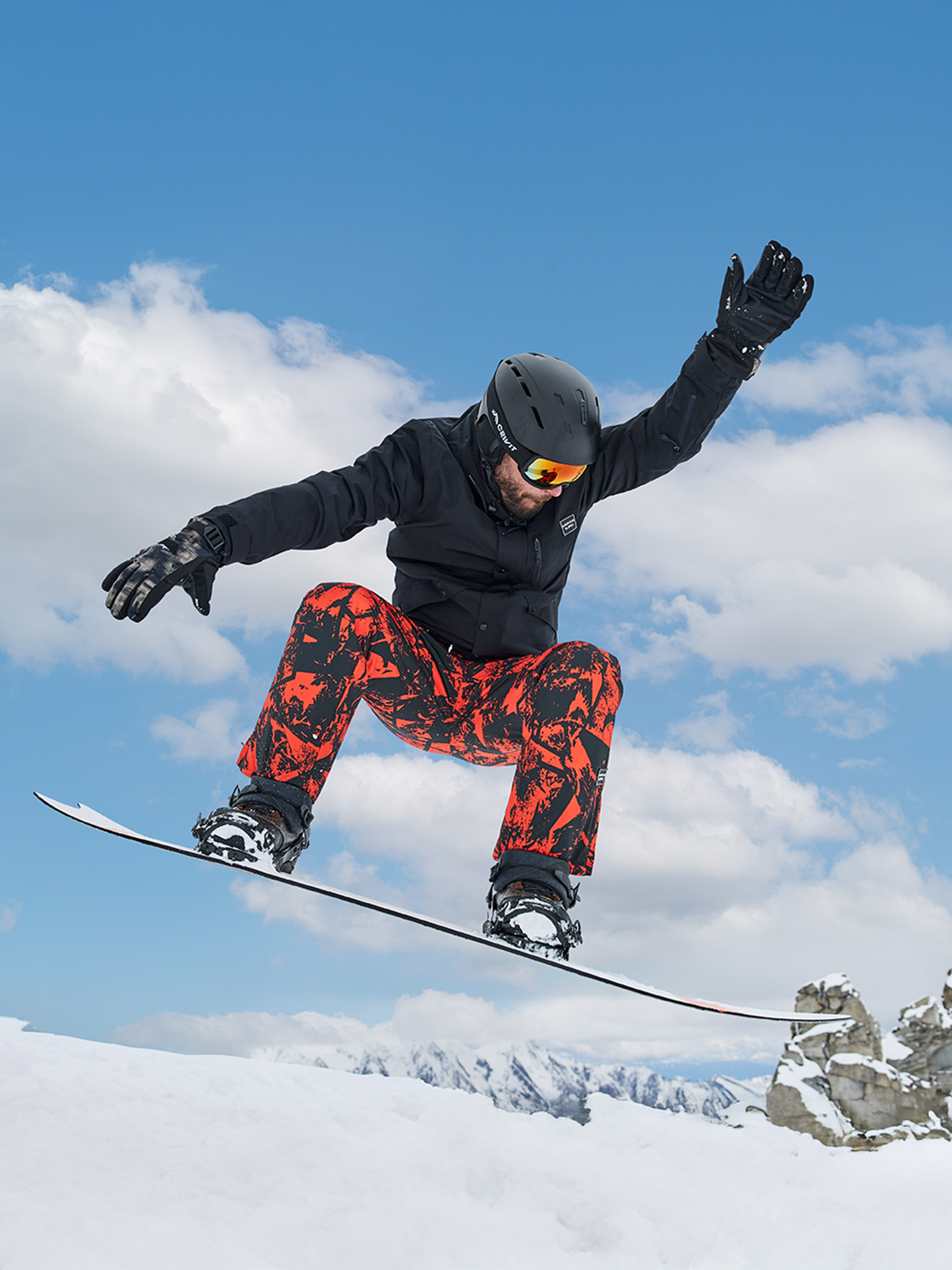 Snowboarder in rot-schwarzer Hose, schwarzer Jacke, Helm und Brille, springt auf einem Snowboard.