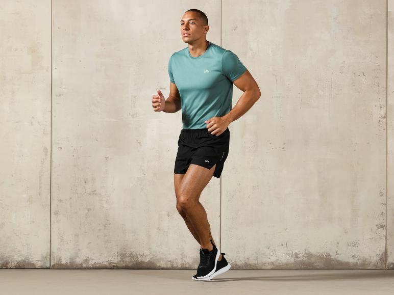 Mann in einem blauen T-Shirt und schwarzen Shorts, posiert vor einer Betonwand.