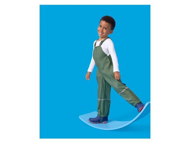 Junge in grüner Latzhose und blauen Stiefeln steht auf einem blauen Balancebrett.