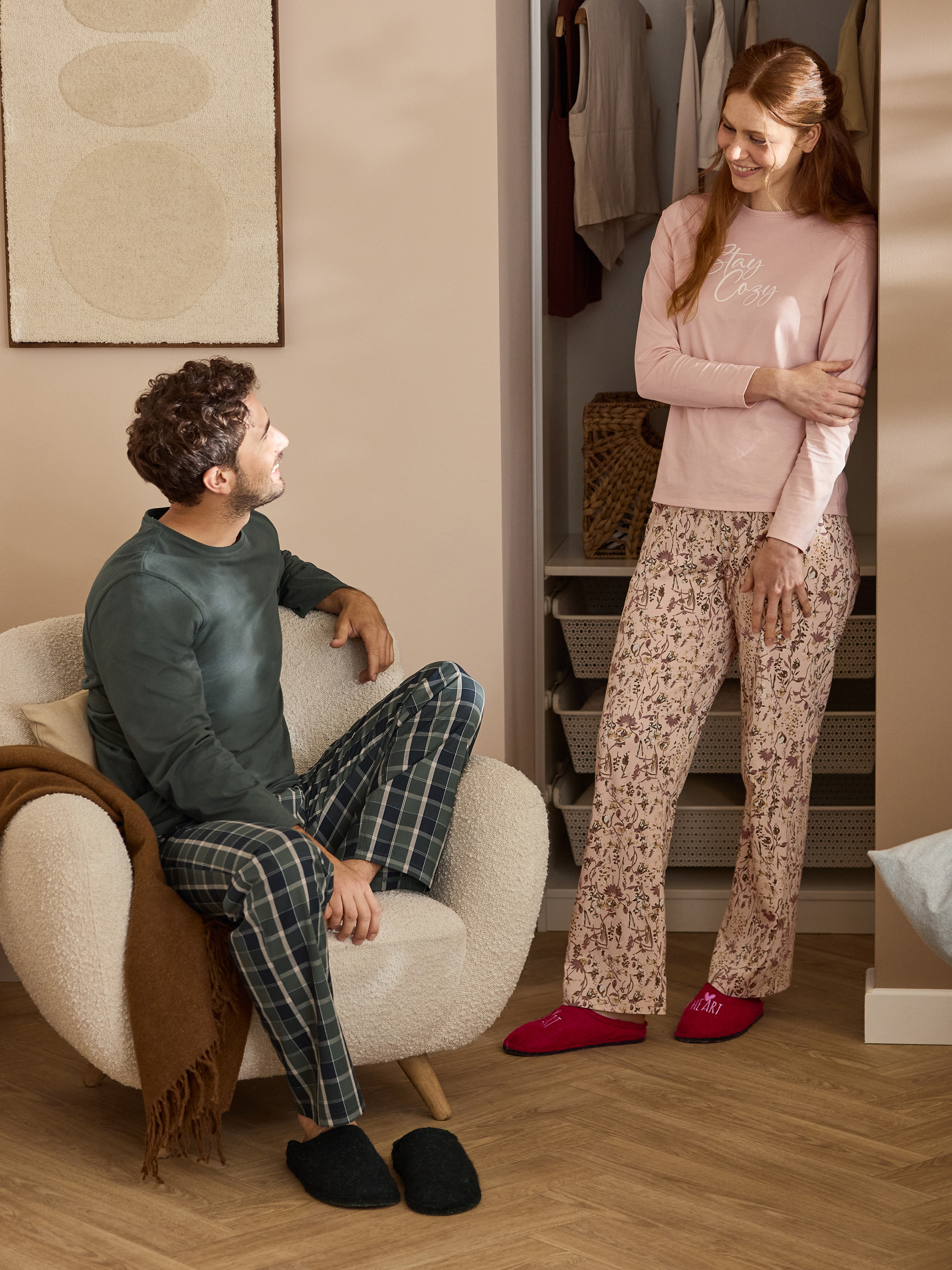Paar in Pyjamas und Hausschuhen, Frau trägt Oberteil mit „Stay Cozy“-Aufschrift.