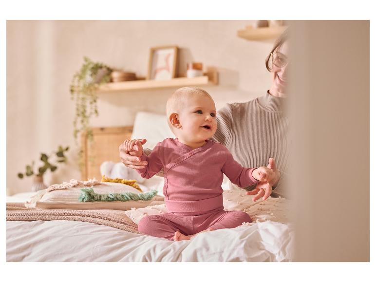 Baby in rosa Kleidung sitzt mit einem Erwachsenen auf einem Bett.