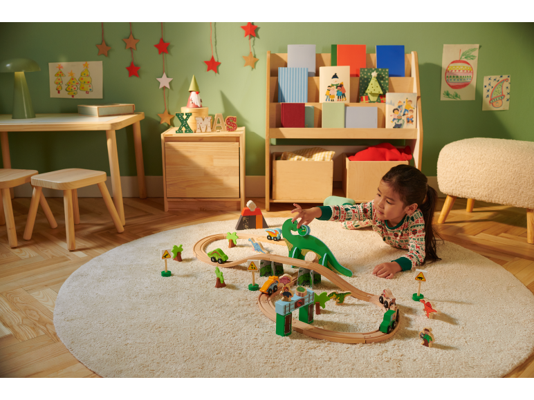 Mädchen spielt mit Holzeisenbahn und Dinosauriern auf einem runden Teppich in einem weihnachtlich dekorierten Zimmer.