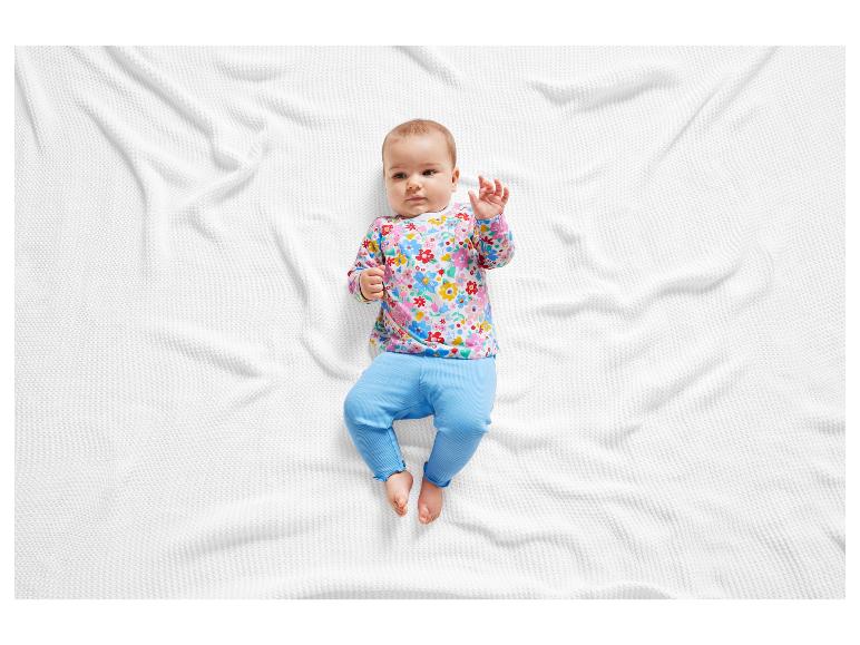 Baby in einem rosafarbenen Blümchenoberteil und blauen Hosen auf einem weißen Bettlaken.