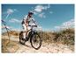 Lächelnder Mann auf E-Bike auf Sandweg mit hohem Gras unter blauem Himmel.