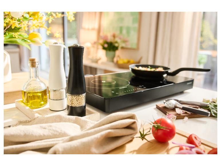 Induktionskochfeld mit Pfanne, Gewürzmühlen, Olivenöl und frischem Gemüse auf einer Küchenarbeitsplatte.