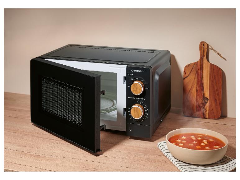 Schwarze Mikrowelle mit offener Tür, daneben eine Schale Tomatensuppe und ein Holzschneidebrett.