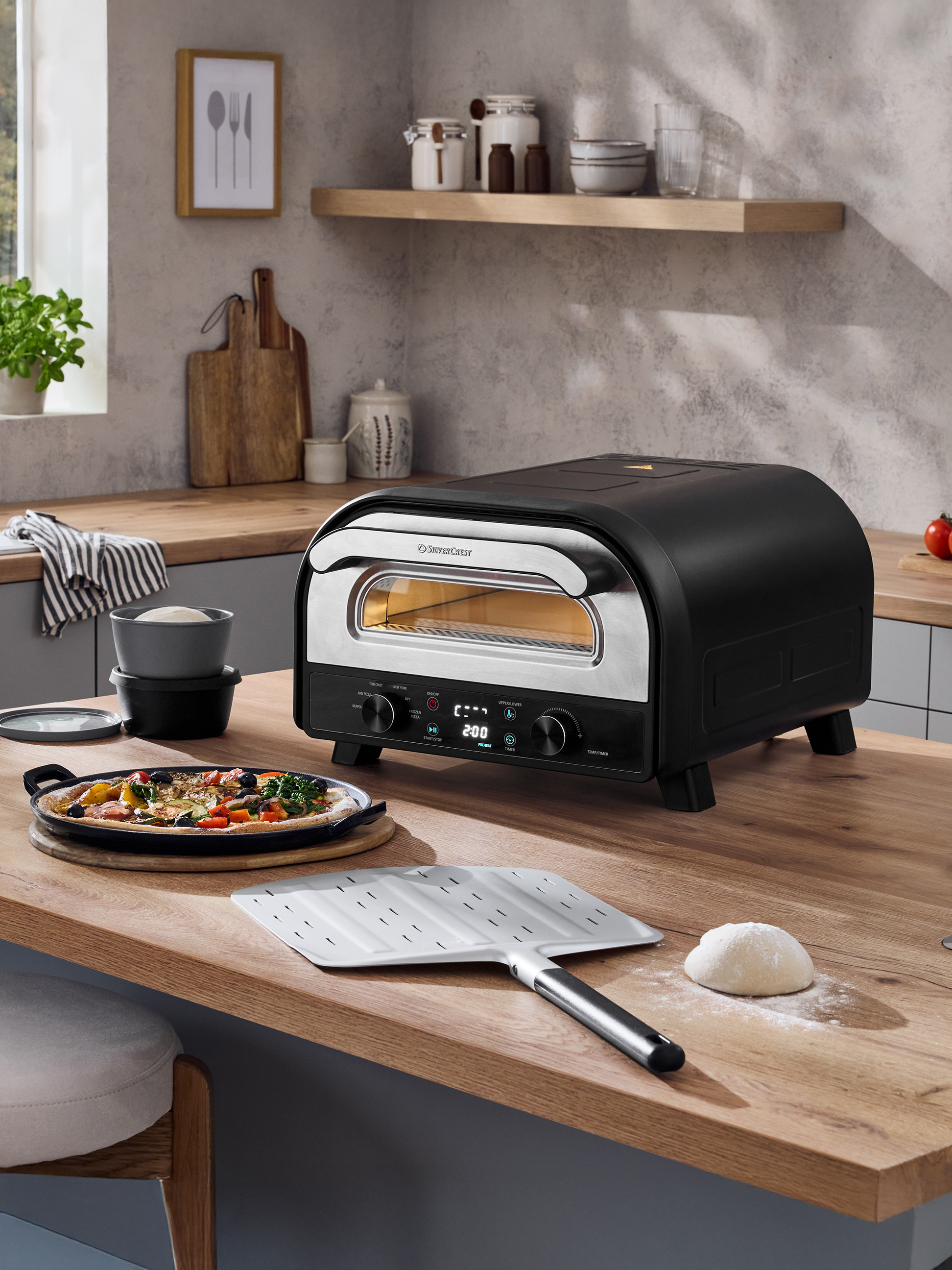 Schwarzer Pizzaofen auf einer Holztheke mit frisch zubereiteter Pizza und Teig.