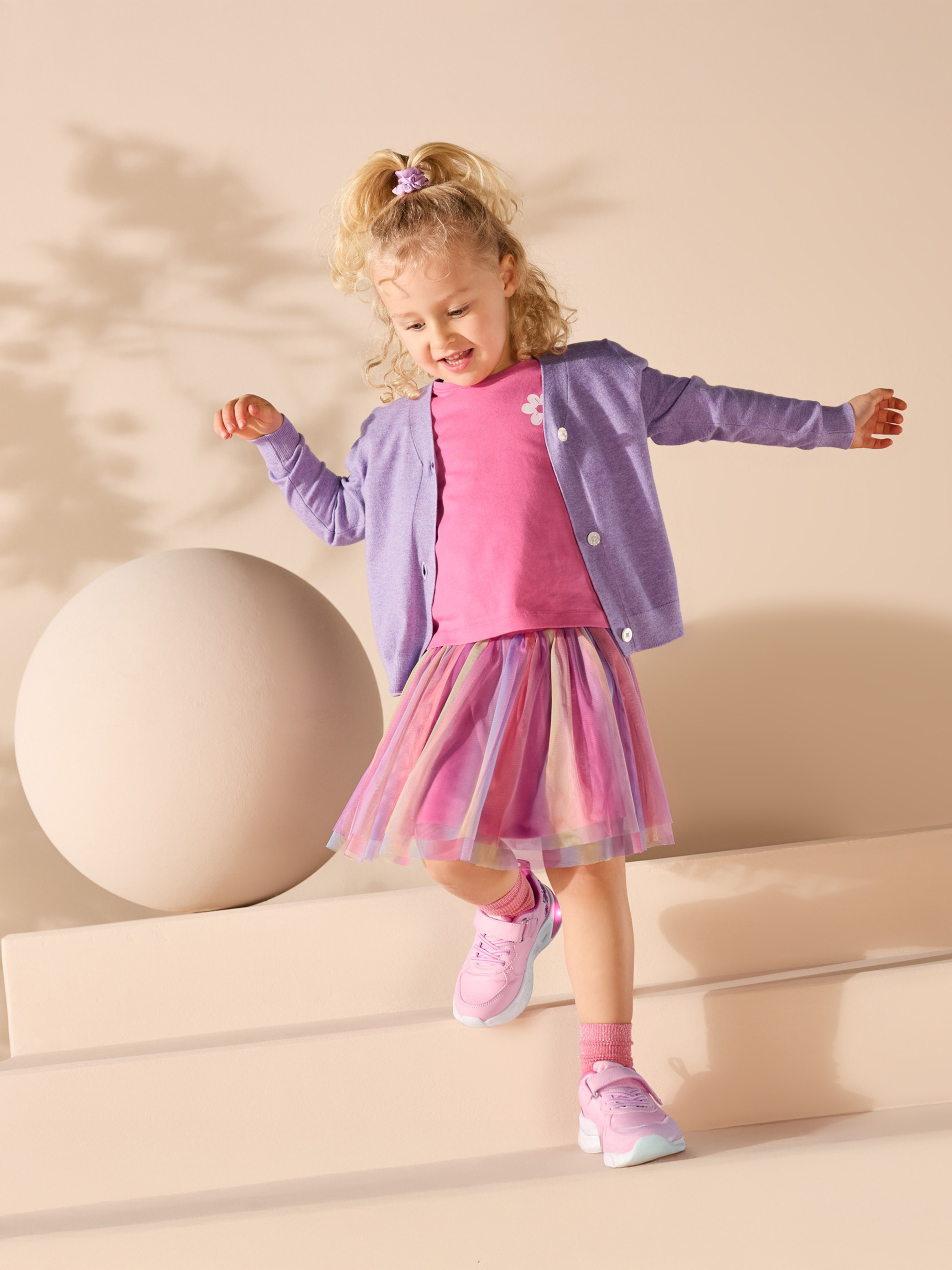 Mädchen in rosa T-Shirt, lila Strickjacke und buntem Tüllrock.