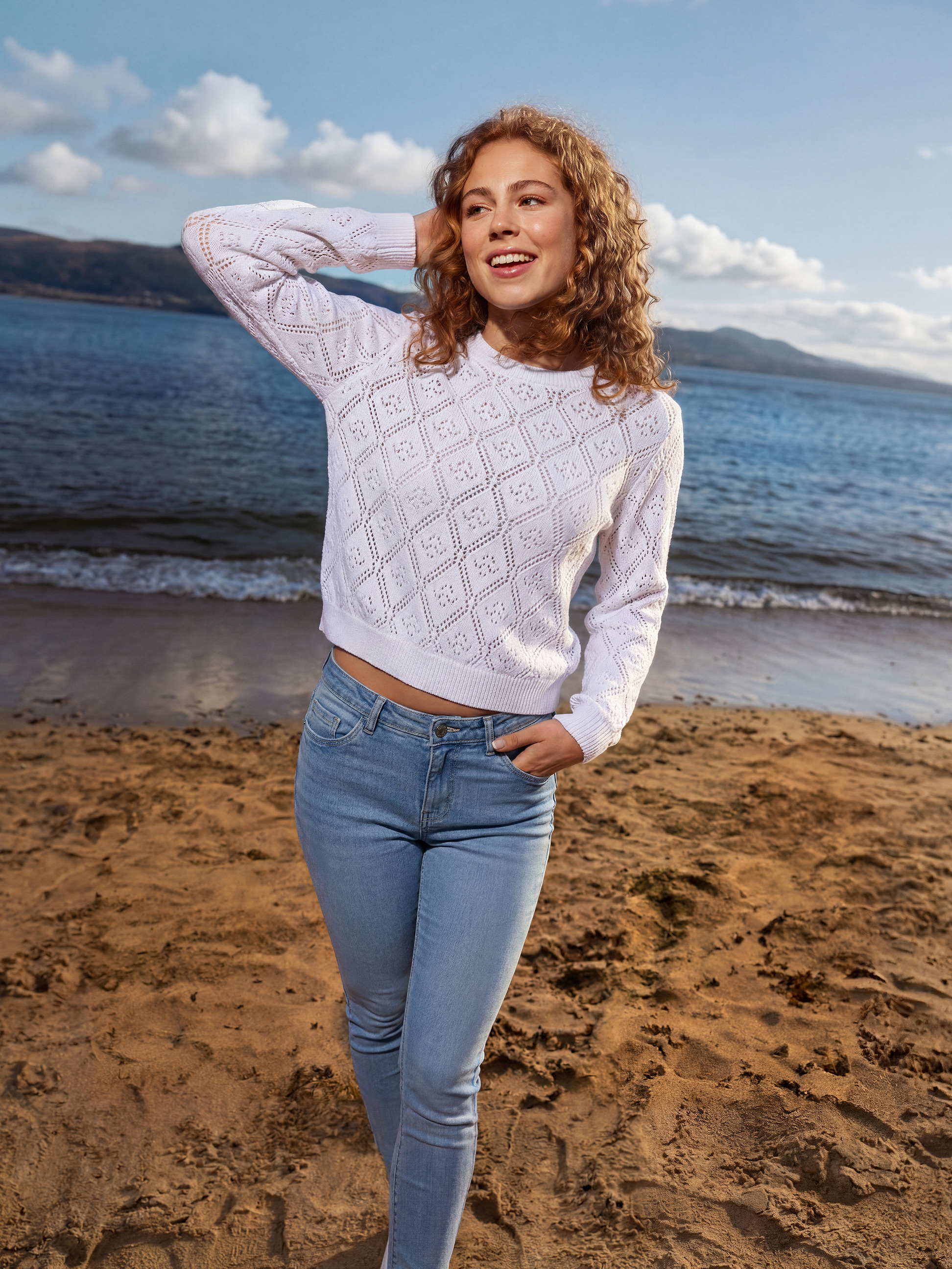 Frau in weißem Pullover und Jeans am Strand, mit Meer und Bergen im Hintergrund.