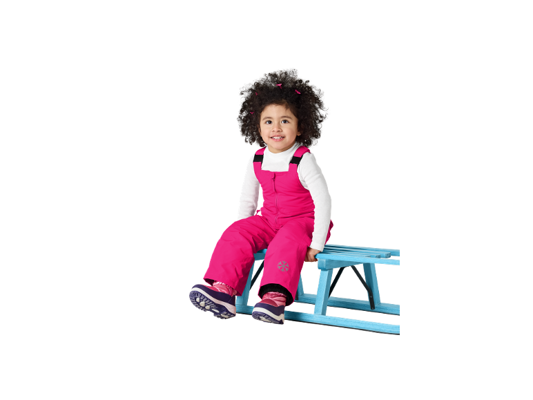Lächelndes Kleinkind in rosa Schneeanzug und Winterstiefeln, sitzend auf einem blauen Schlitten.