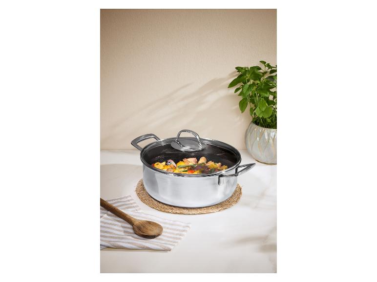 Edelstahltopf mit Glasdeckel, gefüllt mit Gemüse und Fleisch, auf einem Stroh-Tischset.
