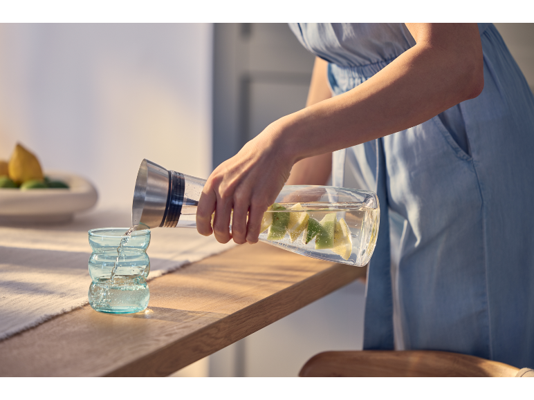 Frau gießt Limettenwasser aus Karaffe in Glas auf Holztisch.