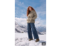 Frau in beiger Pufferjacke, weiten Jeans und Stiefeln steht in verschneiter Berglandschaft.