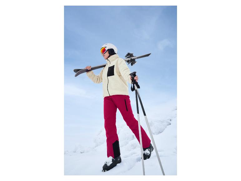 Eine Frau in roten Skianzügen und einem weißen Crivit-Helm geht mit Skiausrüstung durch den Schnee.