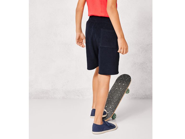 Ein Junge trägt blaue Shorts, Sneakers und ein Skateboard.