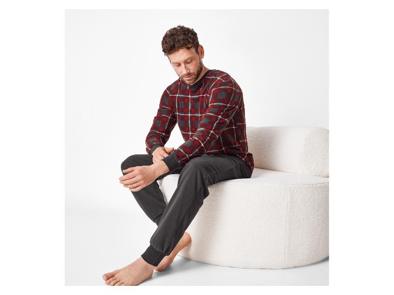 Mann in rotem kariertem Pyjama und grauer Hose, sitzt auf einem weißen Sofa.