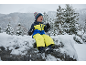 Kind in gelb-blauem Skianzug, Mütze und Handschuhen, sitzt auf einem verschneiten Felsen.