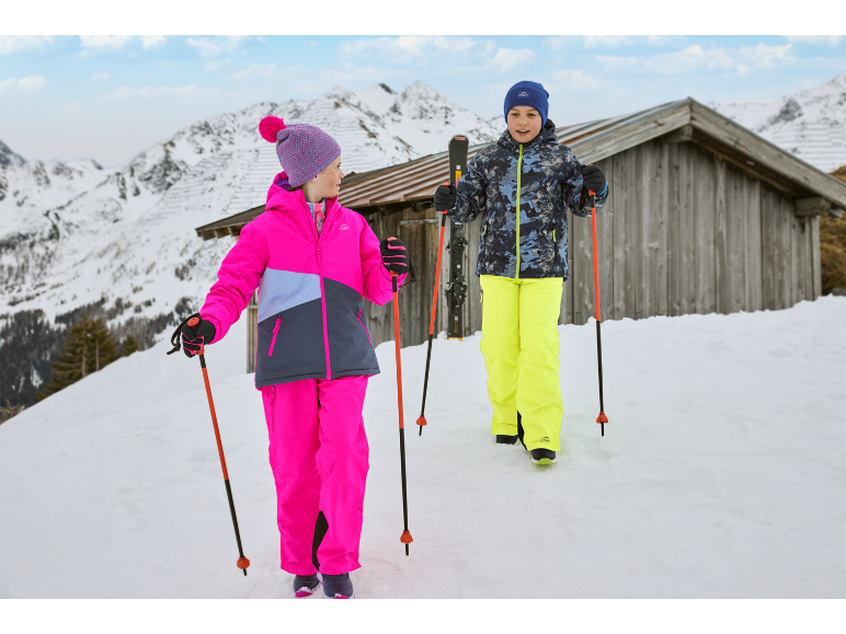 Zwei Kinder in bunter Skibekleidung mit Skistöcken gehen im Schnee neben einer Hütte.