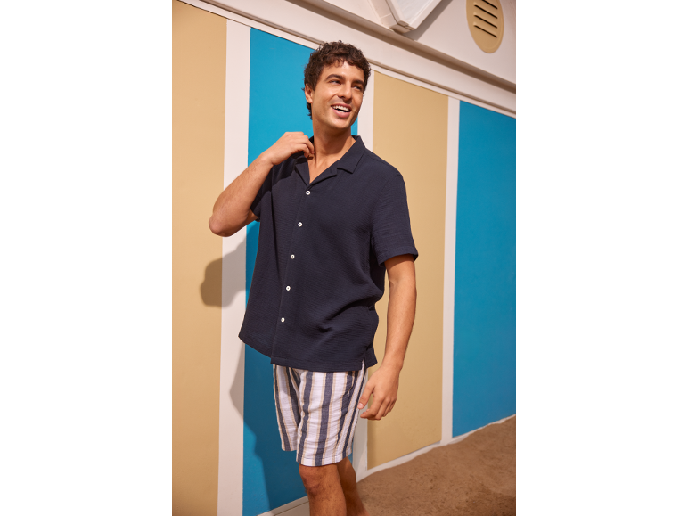 Mann in einem marineblauen Kurzarmhemd und gestreiften Shorts, lächelnd in einer Strandhütten-Umgebung.