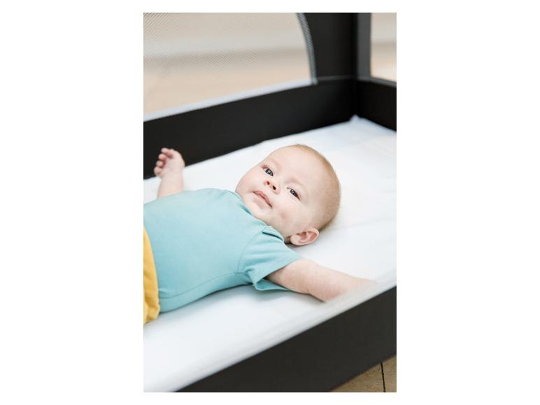 Baby in blauem Body und gelber Hose liegt in einem Reisebett.