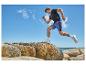 Mann läuft in schwarzem T-Shirt und blauen Shorts auf Felsen, mit Meer und blauem Himmel im Hintergrund.