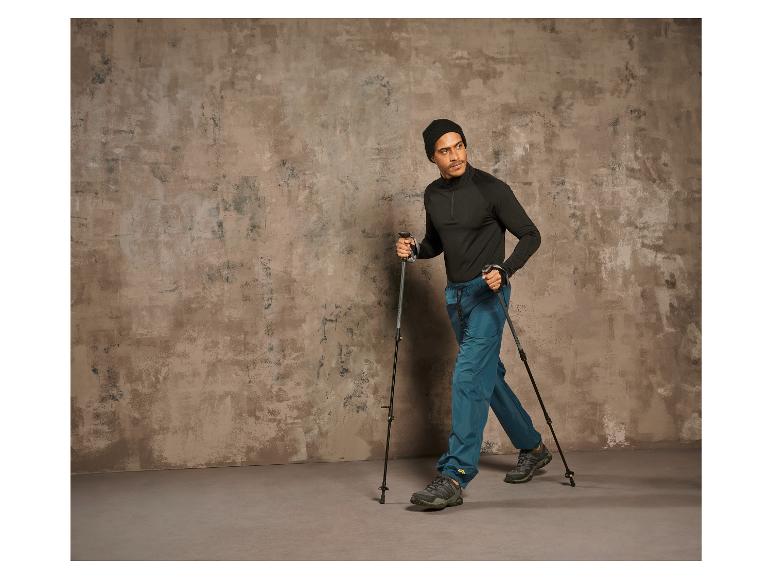 Mann mit Wanderstöcken und Wanderschuhen geht vor einer Betonwand.