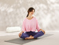 Eine Frau sitzt im Lotussitz auf einer Yogamatte mit einem Meditationskissen daneben.