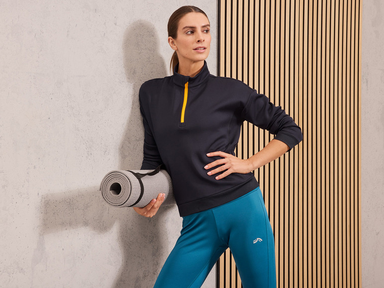 Eine Frau in schwarzer Sportbekleidung und blauen Leggings hält eine Yogamatte.